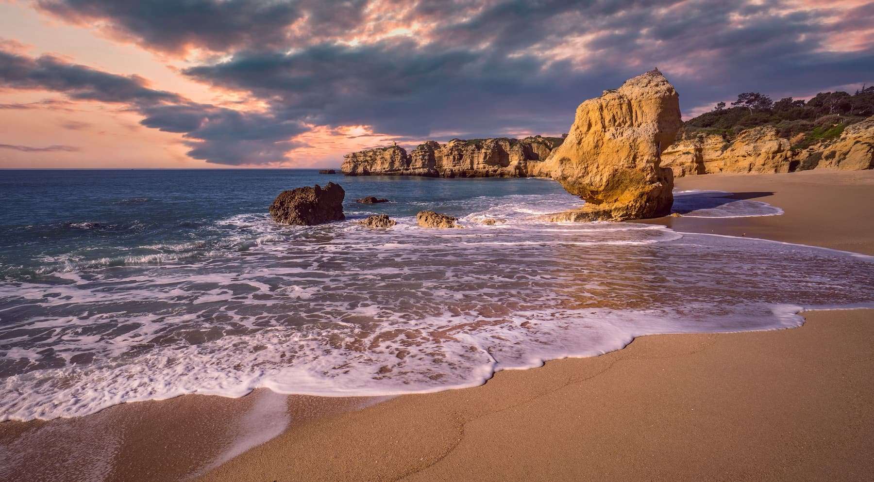 Waves washing a sandy Algarve beach below ochre cliffs and sea stacks at dusk