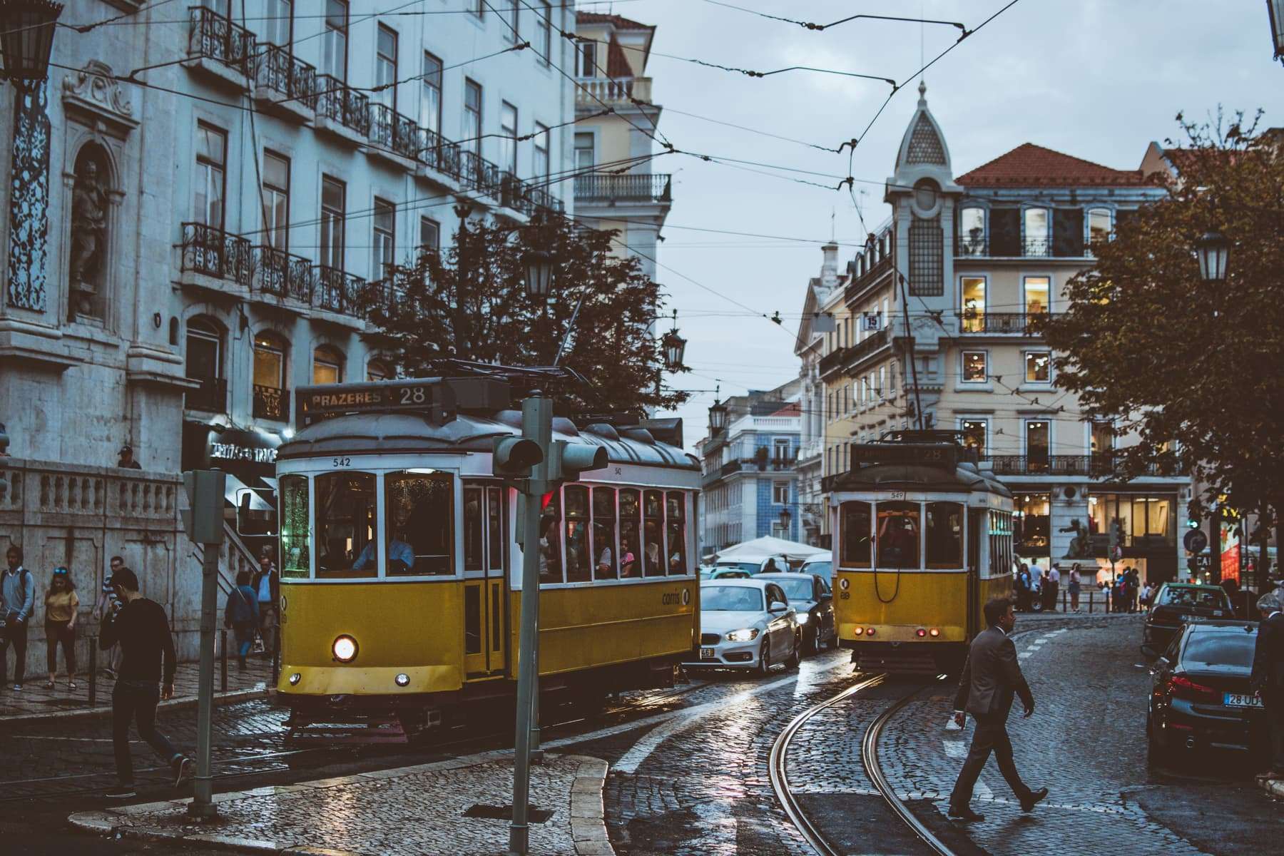 Two yellow number 28 trams and cars on a wet cobbled street in central Lisbon