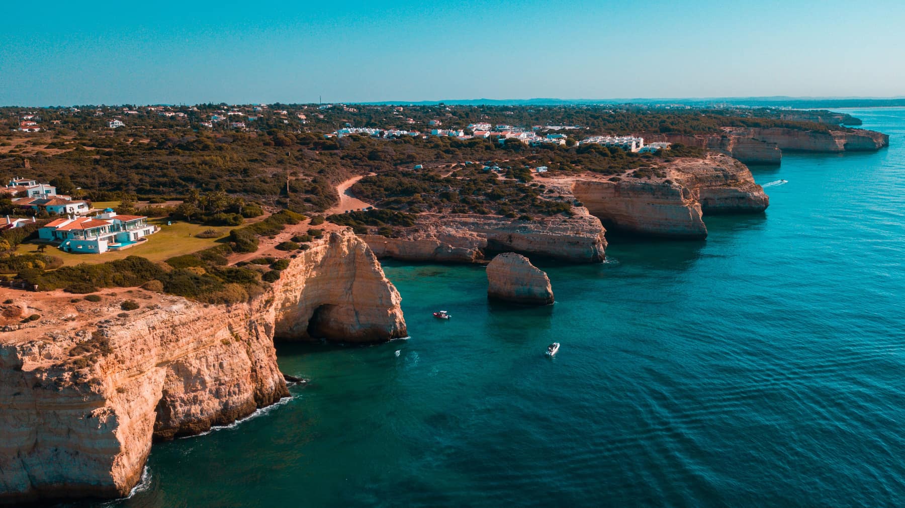 Aerial view of the Algarve cliff coast with sea stacks, boats and clifftop houses reached by a winding track